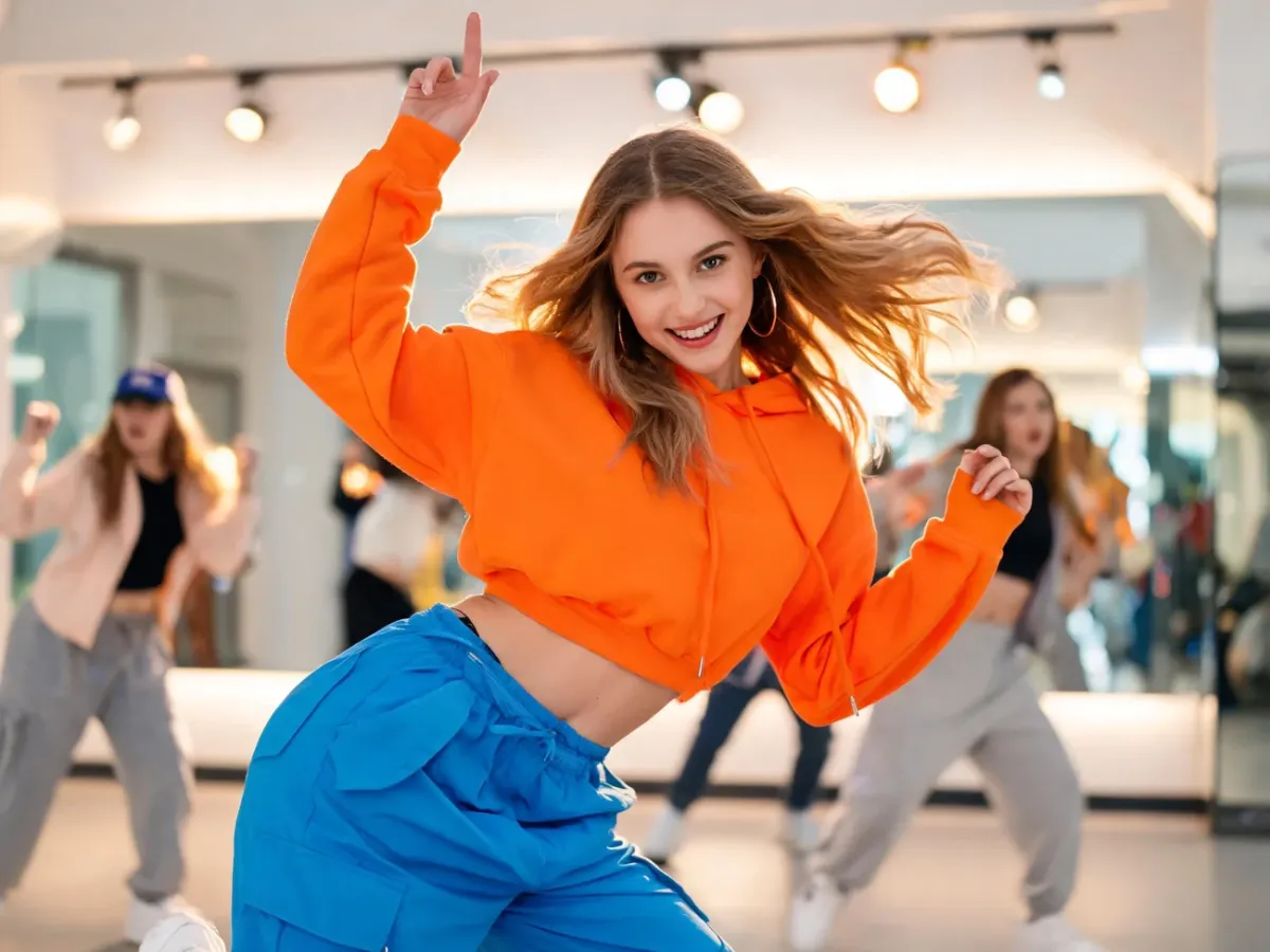 Tancerka w stylu dancehall w dynamicznej pozie w jasnym studio, w tle inni tancerze i lustra podkreślające energię tańca.