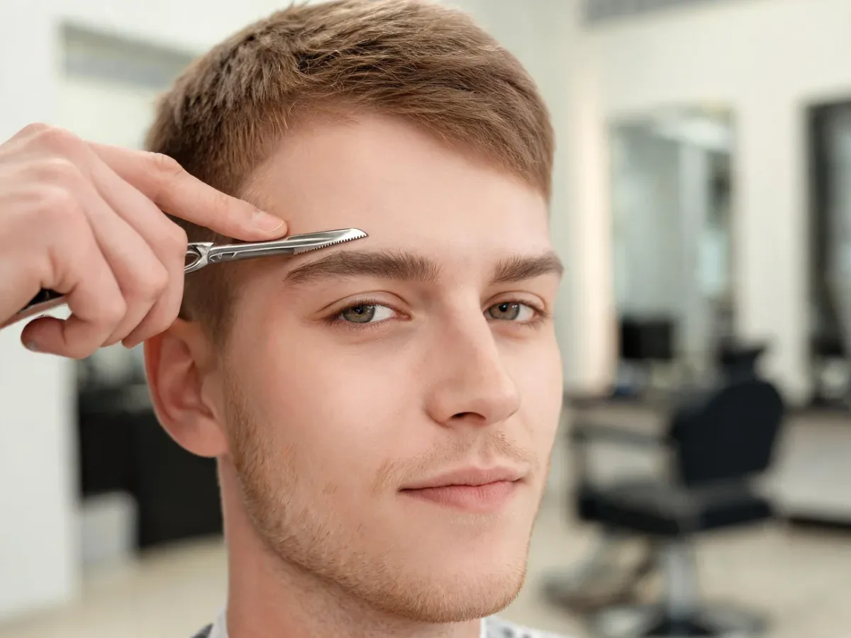 Młody mężczyzna w barber shopie, barber wykonuje precyzyjne nacięcie brwi brzytwą, spokojny, pewny wyraz twarzy.